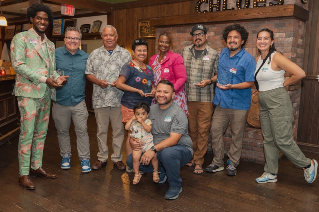 Juan D. Martinez Pineda kneeling with his daughter in front of a group of male and female Black, Brown and white leaders. Many of them, including his wife standing behind him are holding awards.